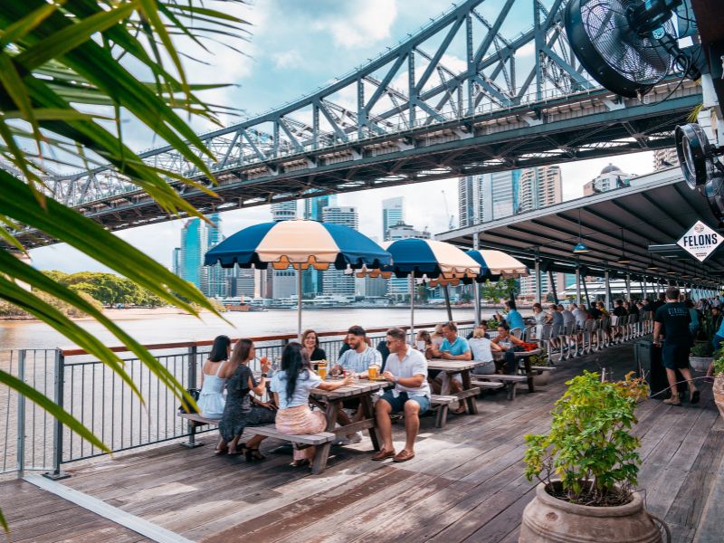 A group of people sat at howard smith wharves enjoying food and drink along the river.