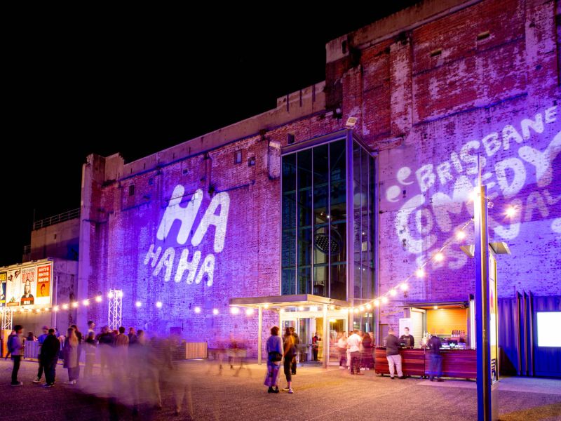 Brisbane Comedy Festival 2026 An exterior shot of Brisbane Powerhouse with purple projections of the comedy festival logo on the walls. People walking around and entering.